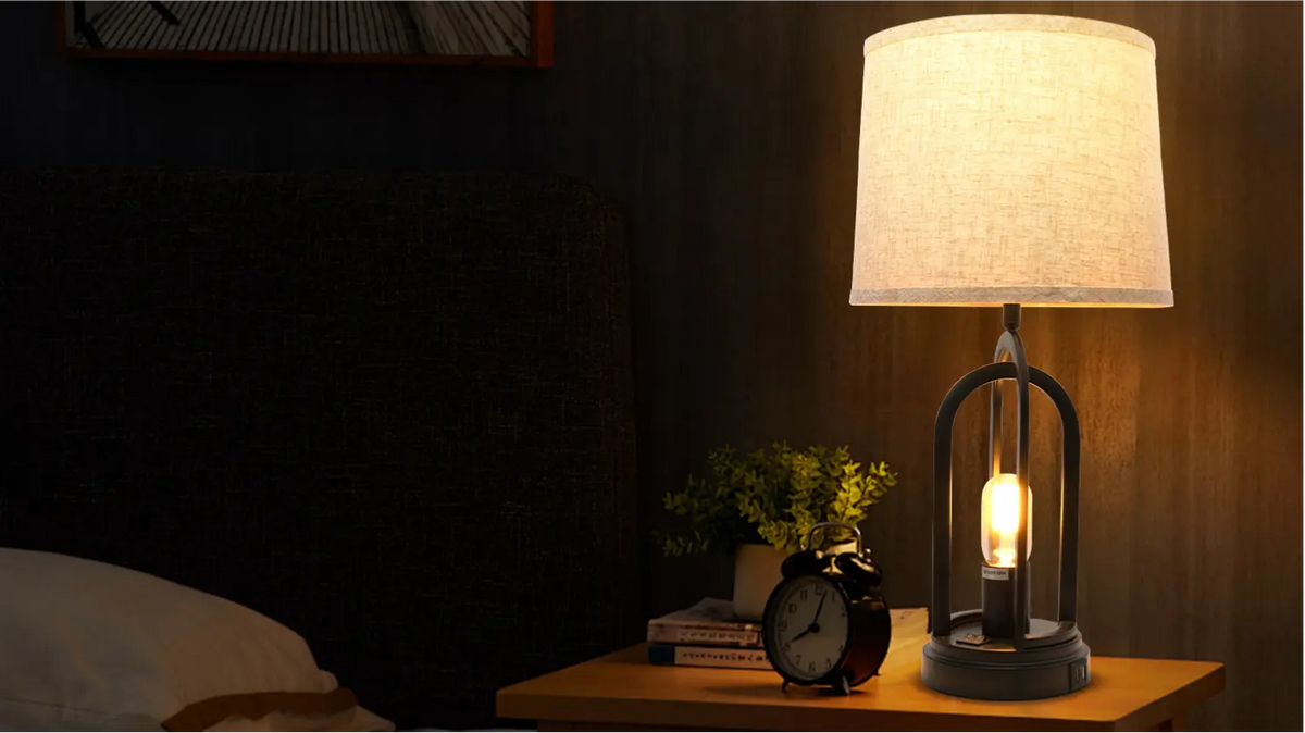 Linen shade table lamp with a round metal cage base glowing warmly on a wooden nightstand beside a bed and alarm clock