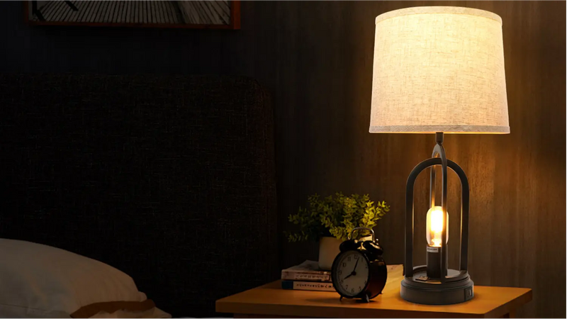 Linen shade table lamp with a round metal cage base glowing warmly on a wooden nightstand beside a bed and alarm clock