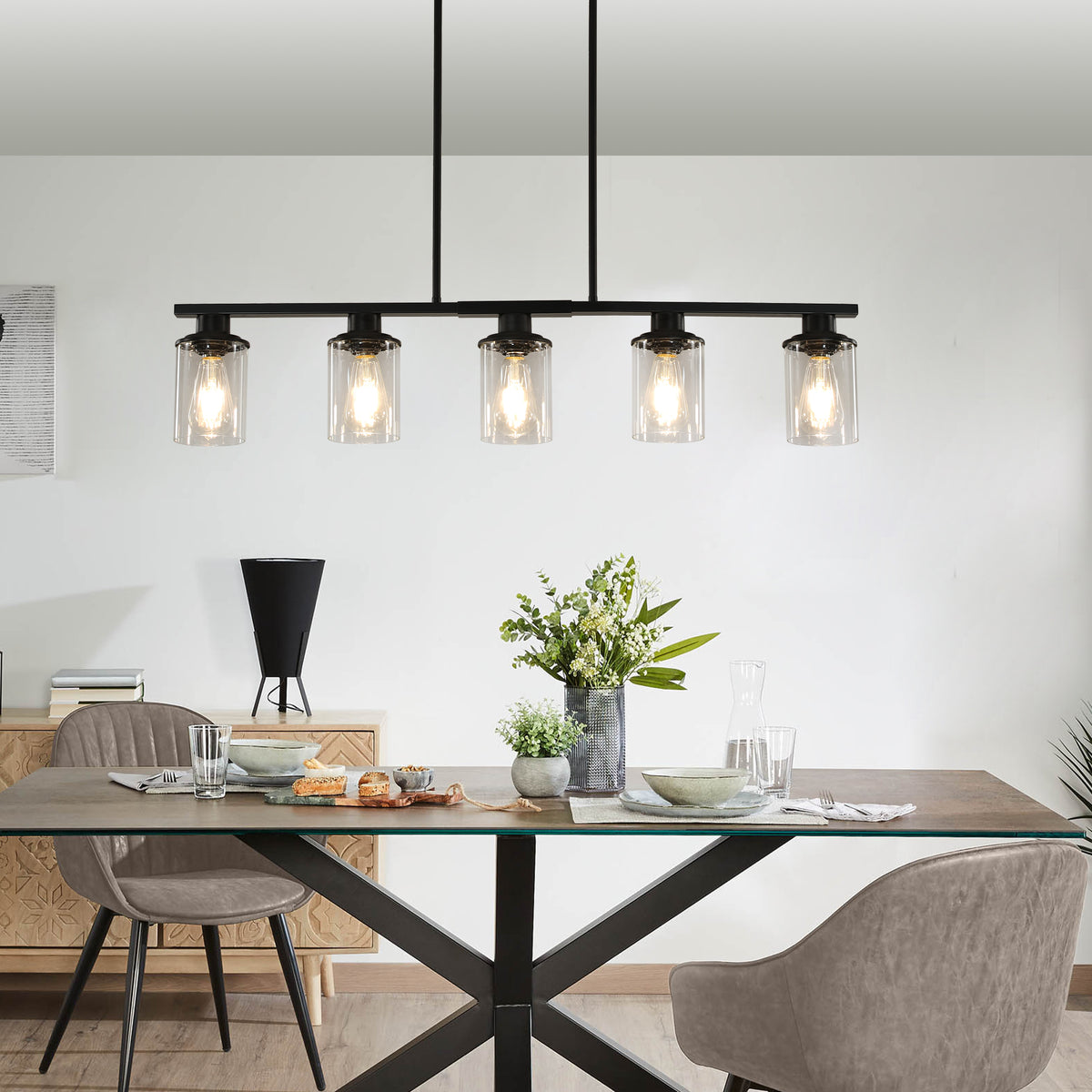 Black Ashcombe Glass Shade Chandelier hanging above a wooden dining table, featuring a linear five-light bar, clear cylindrical glass shades, and three black suspension rods.