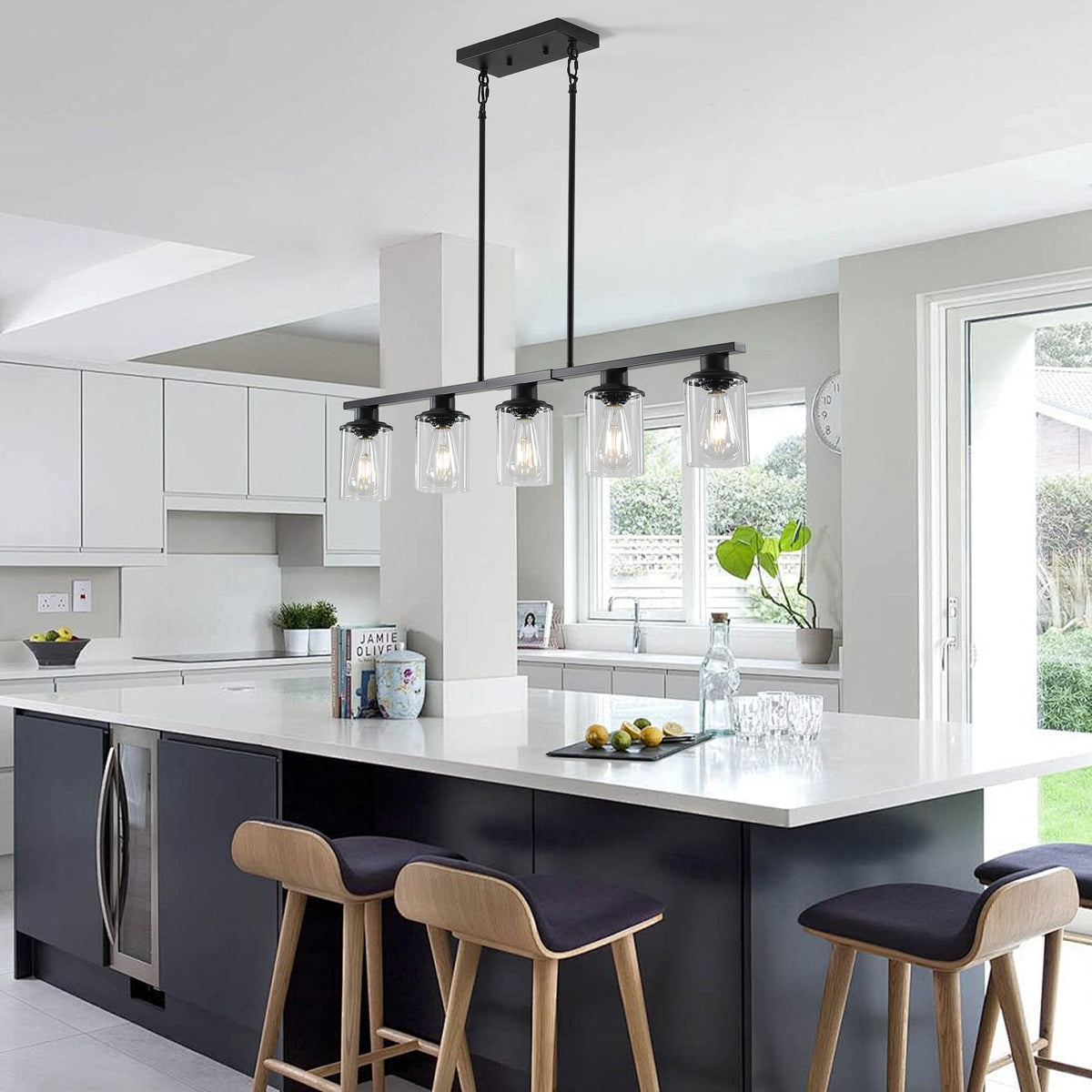 Black Ashcombe Glass Shade Chandelier installed over a white kitchen island, featuring five clear glass cylinder shades on a linear black metal bar with adjustable vertical rods.