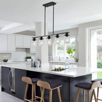 Black Ashcombe Glass Shade Chandelier installed over a white kitchen island, featuring five clear glass cylinder shades on a linear black metal bar with adjustable vertical rods.