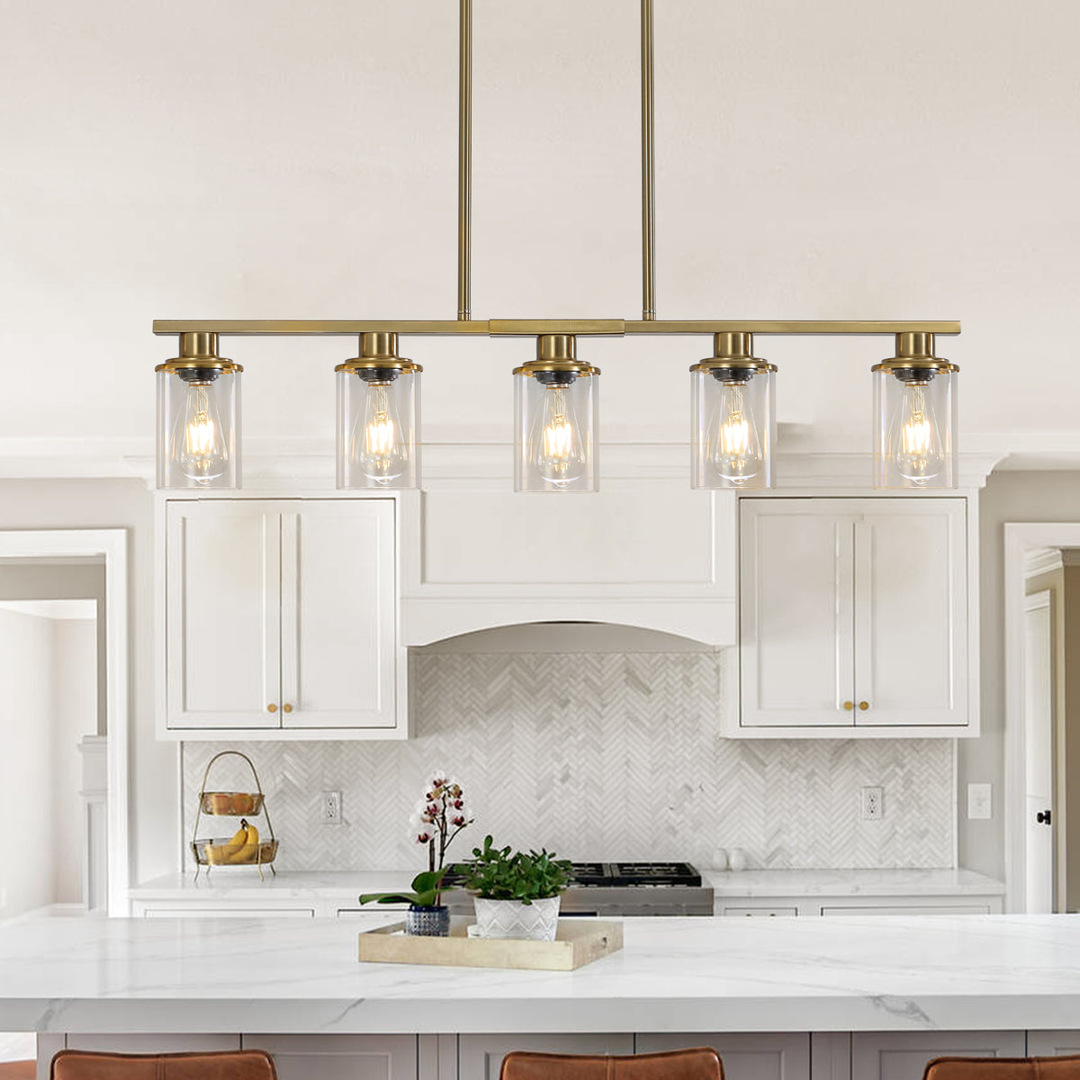 Brass Ashcombe Glass Shade Chandelier installed above a white kitchen island, featuring a linear five-light bar, clear cylindrical glass shades, and slim brass hanging rods.