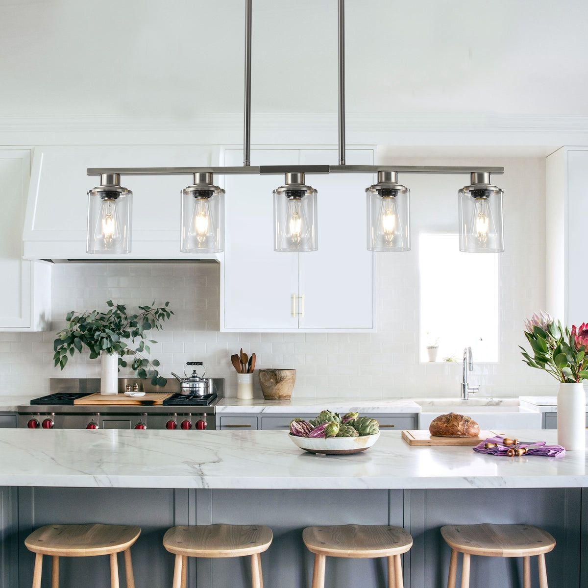 Black Ashcombe Glass Shade Chandelier suspended above a kitchen island with bar stools, featuring five clear glass shades, warm exposed bulbs, and a black linear metal frame.