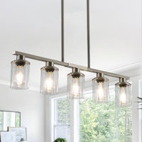 Close-up view of the Ashcombe Glass Shade Chandelier in a brushed metal finish, featuring a horizontal five-light bar, clear cylindrical glass shades, and adjustable hanging rods.